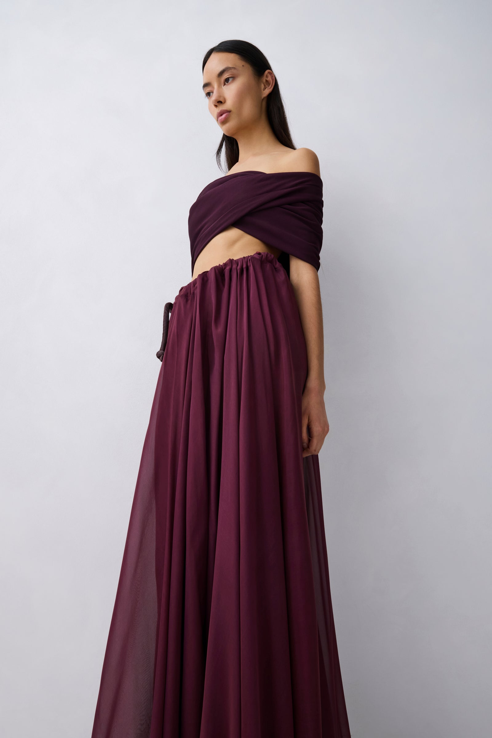 A woman wearing a deep burgundy off-the-shoulder maxi dress with a flowing skirt, shown from the back against a light gray studio background.