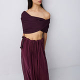 A woman wearing a deep burgundy off-the-shoulder maxi dress with a flowing skirt, shown from the back against a light gray studio background.