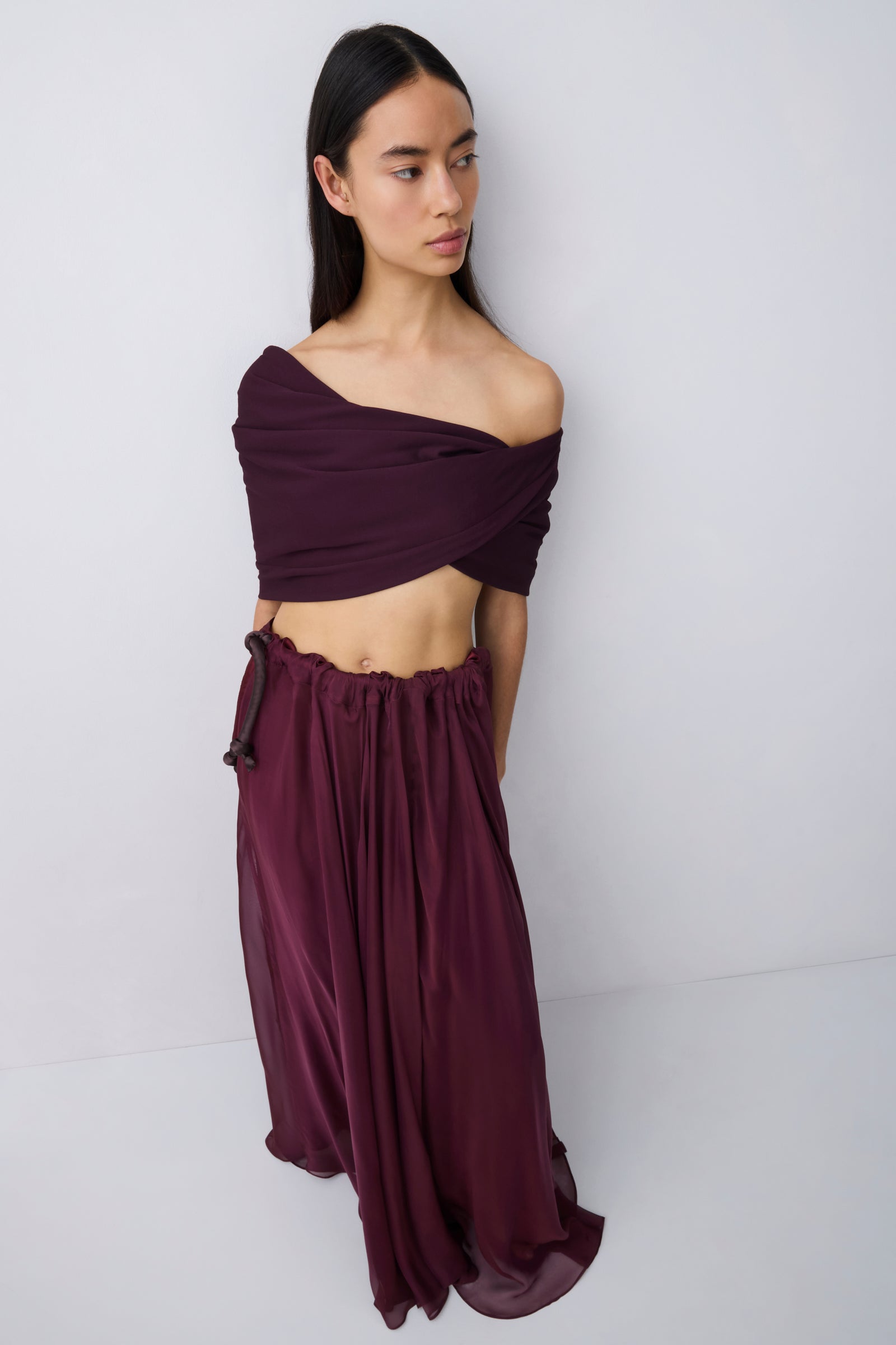 A woman wearing a deep burgundy off-the-shoulder maxi dress with a flowing skirt, shown from the back against a light gray studio background.