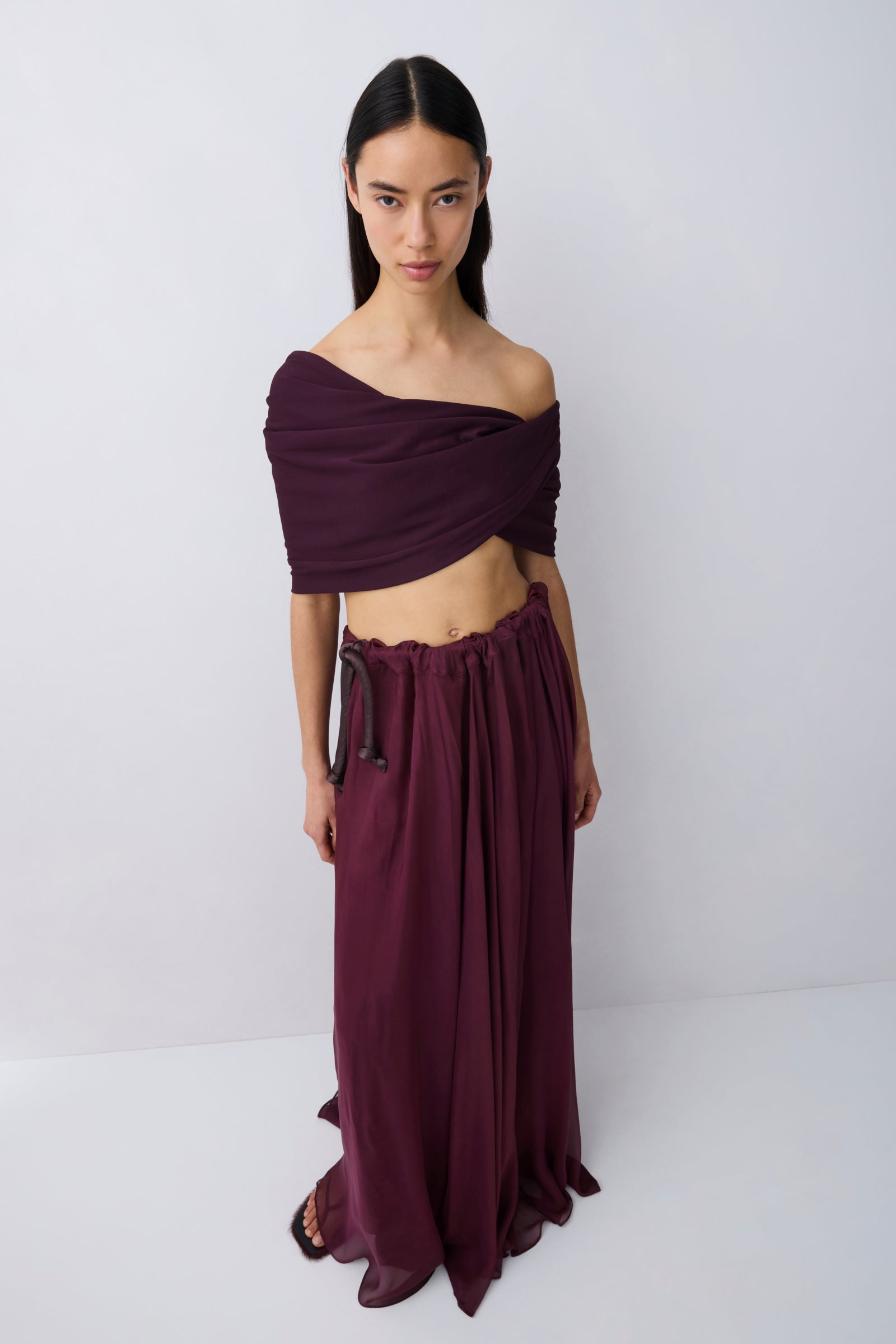 A woman wearing a deep burgundy off-the-shoulder maxi dress with a flowing skirt, shown from the back against a light gray studio background.