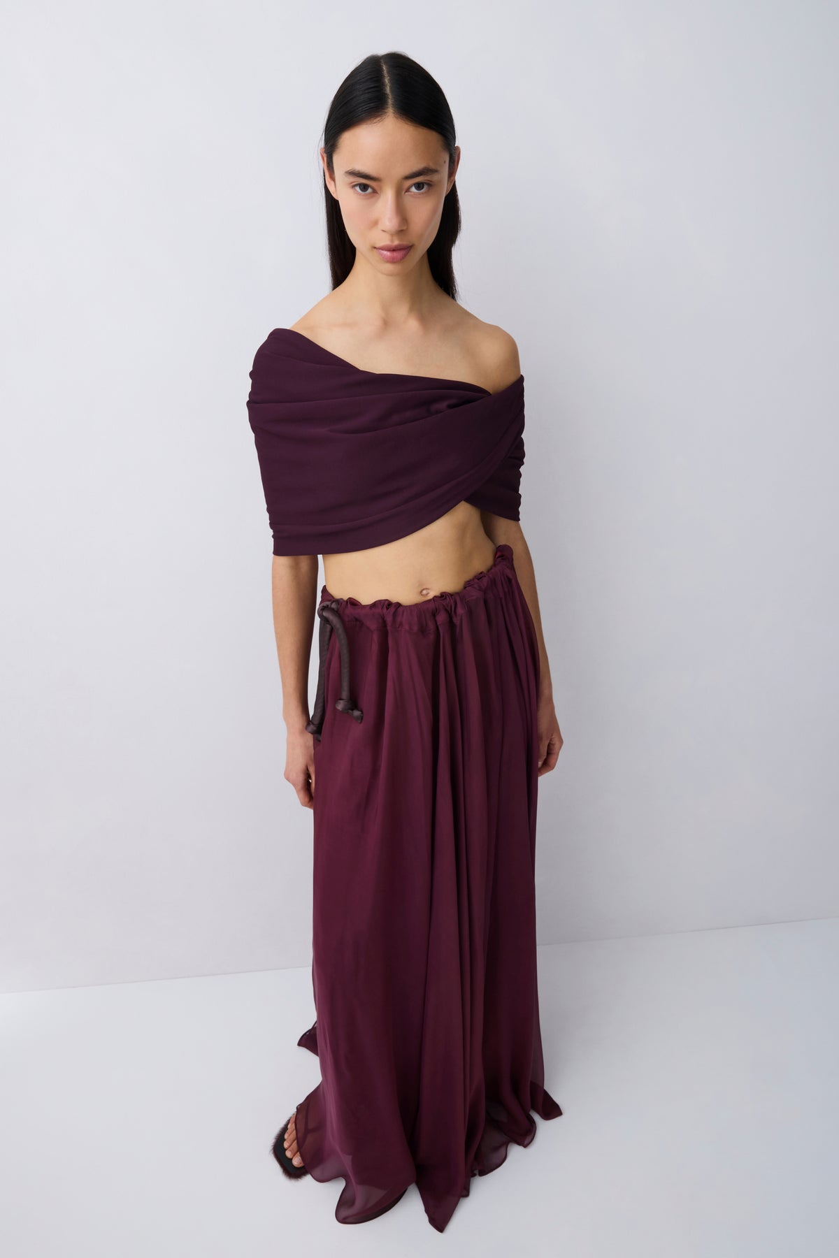A woman wearing a deep burgundy off-the-shoulder maxi dress with a flowing skirt, shown from the back against a light gray studio background.