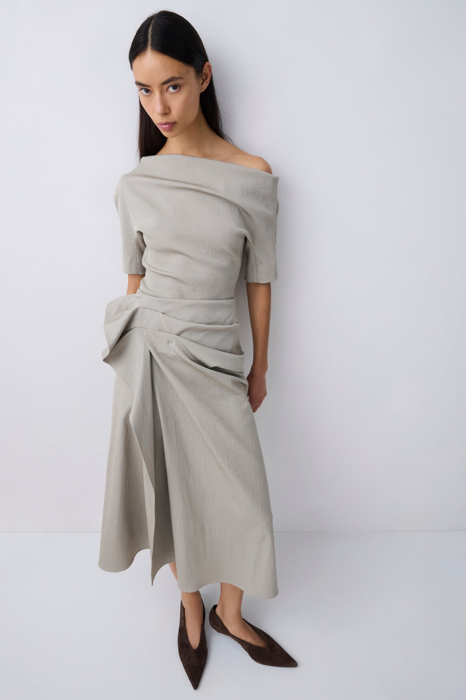 A woman modeling an off-the-shoulder, draped midi skirt in a light taupe-beige color, paired with dark brown pointed-toe flats, against a plain light gray studio background.