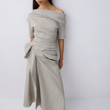 A woman modeling an off-the-shoulder, draped top in a light taupe-beige color, paired with dark brown pointed-toe flats, against a plain light gray studio background.