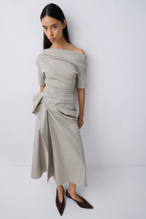 A woman modeling an off-the-shoulder, draped top in a light taupe-beige color, paired with dark brown pointed-toe flats, against a plain light gray studio background.