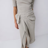 A model wearing a light beige off-the-shoulder knit top  with a draped, gathered skirt and dark brown pointed-toe flats, shown from a side/back angle against a white studio background.