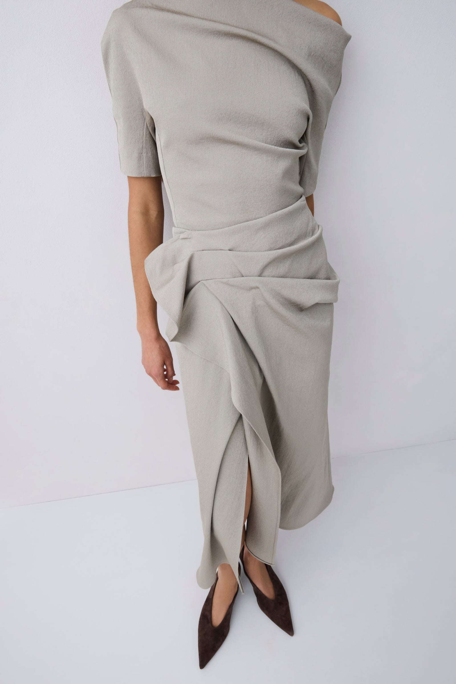 A model wearing a light beige off-the-shoulder knit top  with a draped, gathered skirt and dark brown pointed-toe flats, shown from a side/back angle against a white studio background.
