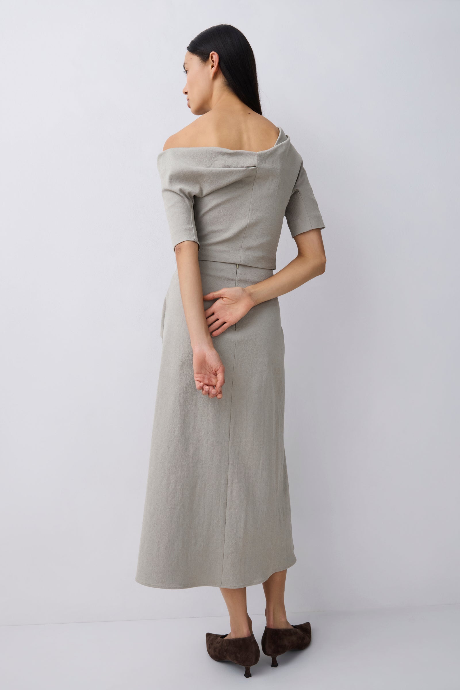A woman modeling an off-the-shoulder top and elevated midi skirt in a light beige linen-like fabric, shown from the back against a plain light gray studio background.