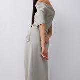 A woman modeling a light gray off-the-shoulder top and midi with short sleeves and a draped neckline and gathered waist, photographed against a plain light gray studio background.