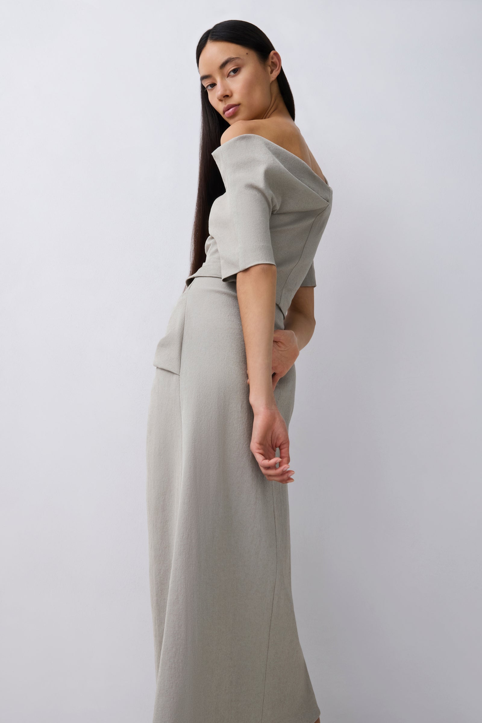 A woman modeling a light gray off-the-shoulder top and midi with short sleeves and a draped neckline and gathered waist, photographed against a plain light gray studio background.