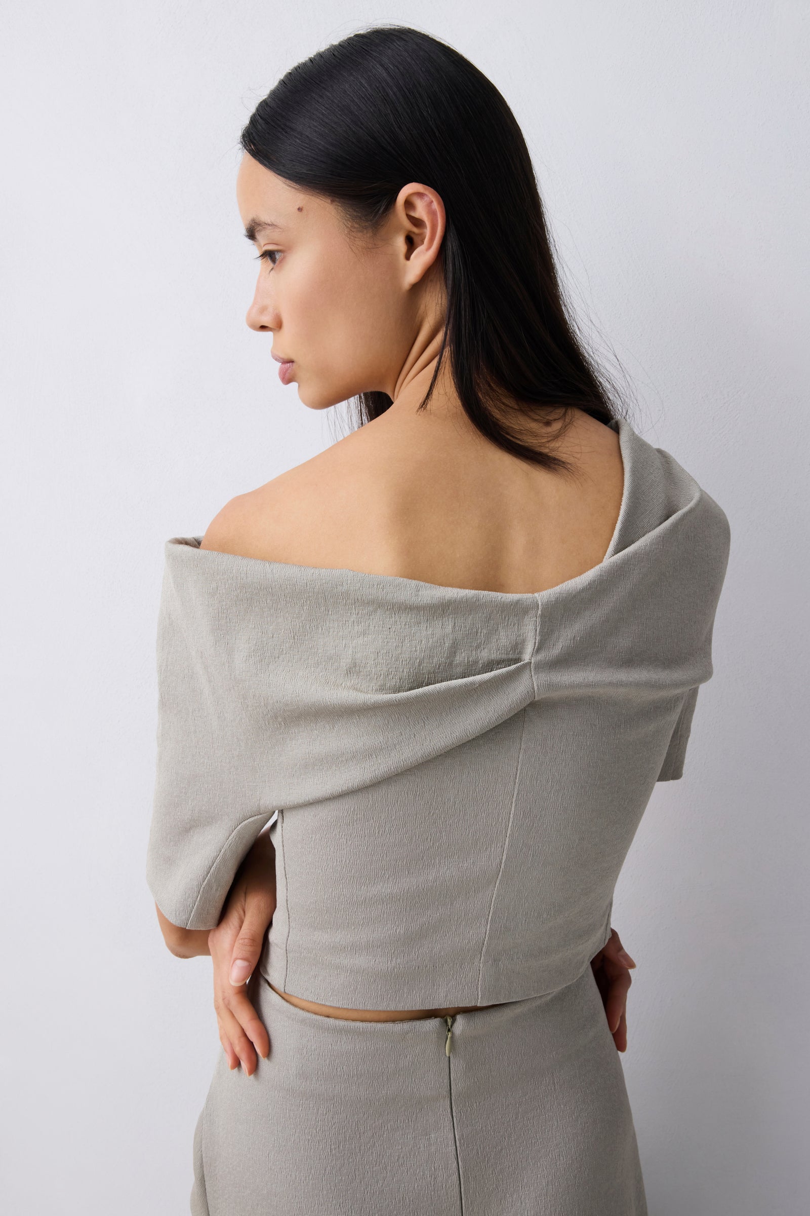 A woman shown from the back wearing a light gray off-the-shoulder linen like top and matching skirt against a plain light gray background.