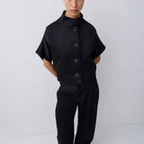 A model stands against a light gray studio background wearing a black short-sleeve button-front top with a high mock neck and black wide-leg trousers.