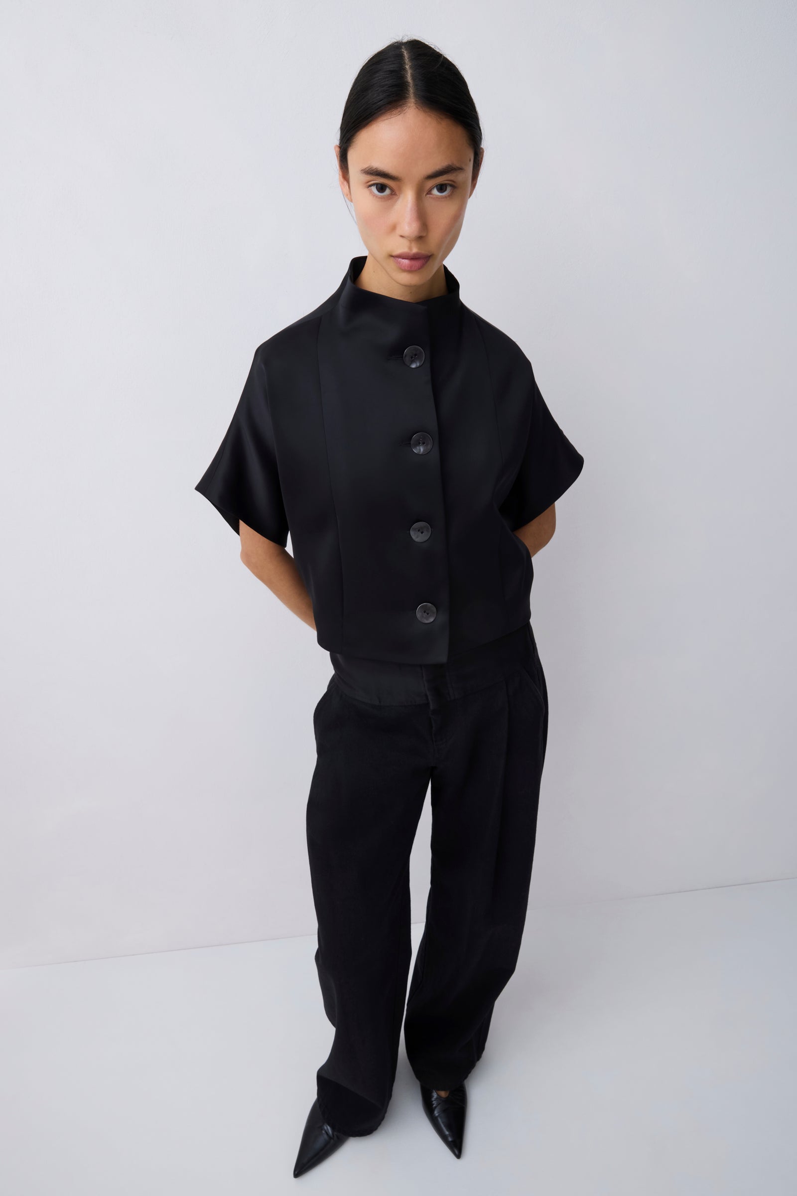 A model stands against a light gray studio background wearing a black short-sleeve button-front top with a high mock neck and black wide-leg trousers.