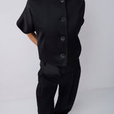 A model wearing a black short-sleeve button-front jacket with a high mock neck, paired with black trousers and pointed black shoes, photographed against a light gray studio background.