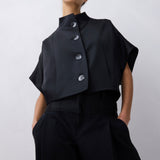 A model wearing a black cropped short-sleeve top with a high mock neck and three prominent silver buttons, paired with black wide-leg trousers, against a light gray studio background.