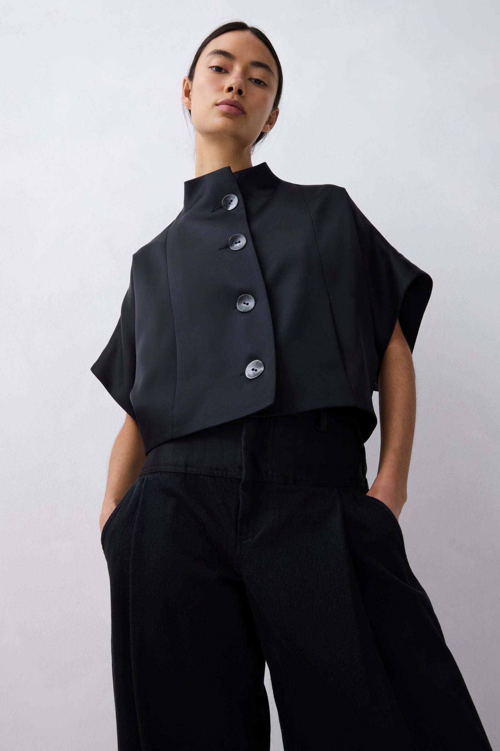 A model wearing a black cropped short-sleeve top with a high mock neck and three prominent silver buttons, paired with black wide-leg trousers, against a light gray studio background.