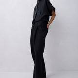 A model wearing a black short-sleeve button-up shirt and matching black wide-leg trousers, standing against a light gray studio background.