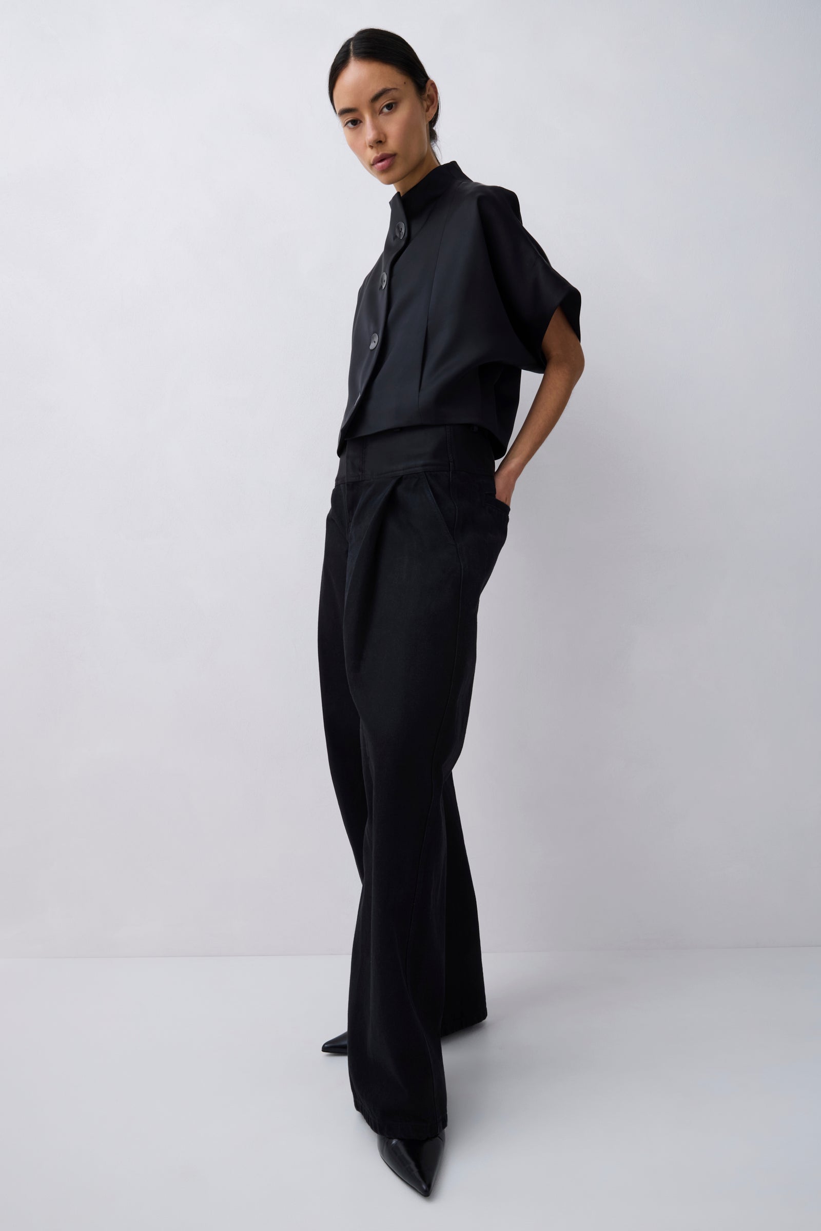 A model wearing a black short-sleeve button-up shirt and matching black wide-leg trousers, standing against a light gray studio background.