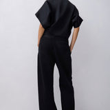 A model viewed from behind wearing a black short-sleeve top with dramatic draped sleeves and matching black wide-leg trousers, set against a plain light gray studio background.