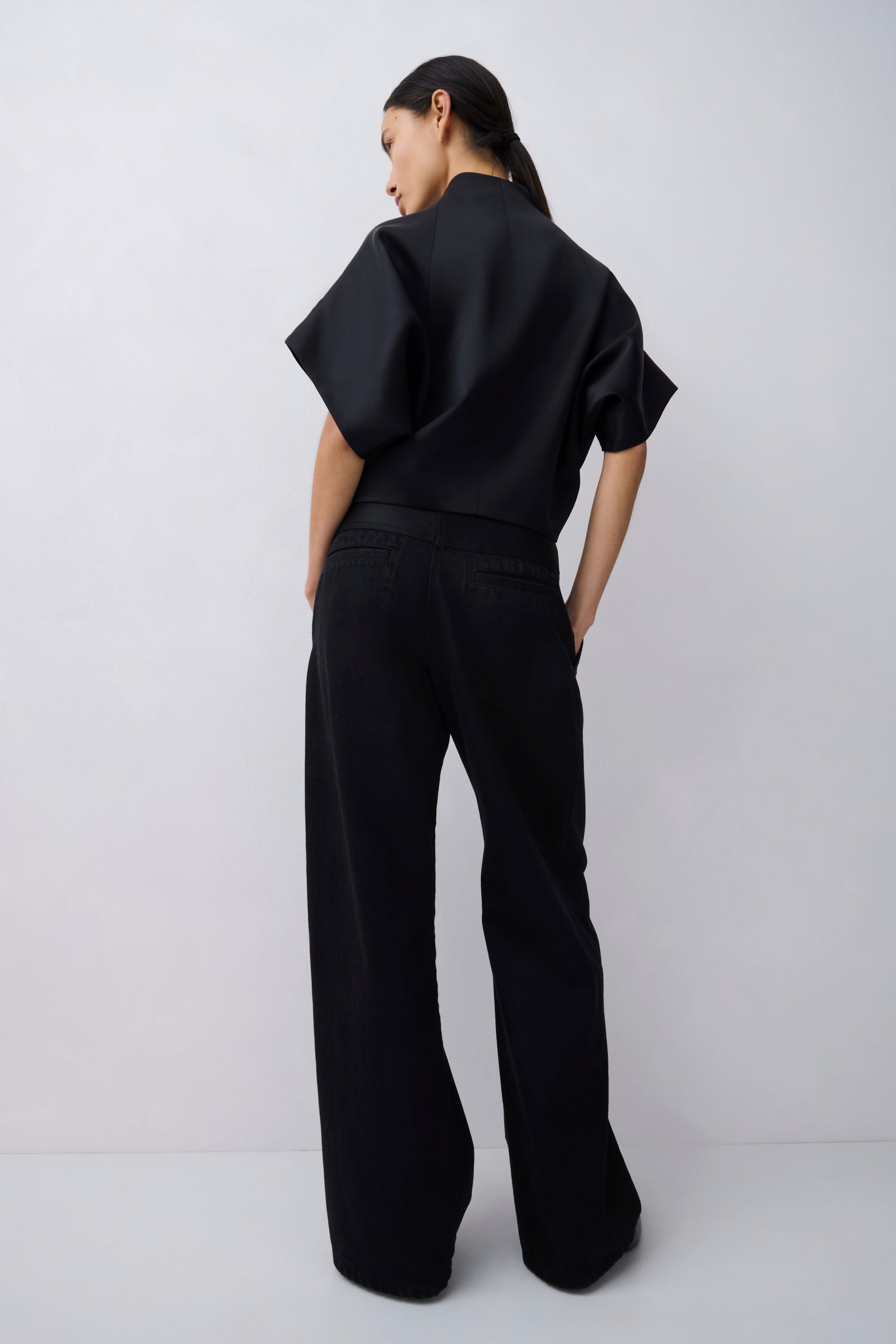 A model viewed from behind wearing a black short-sleeve top with dramatic draped sleeves and matching black wide-leg trousers, set against a plain light gray studio background.