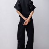 A model viewed from behind wearing a black short-sleeve top and matching wide-leg trousers against a plain light gray studio background.