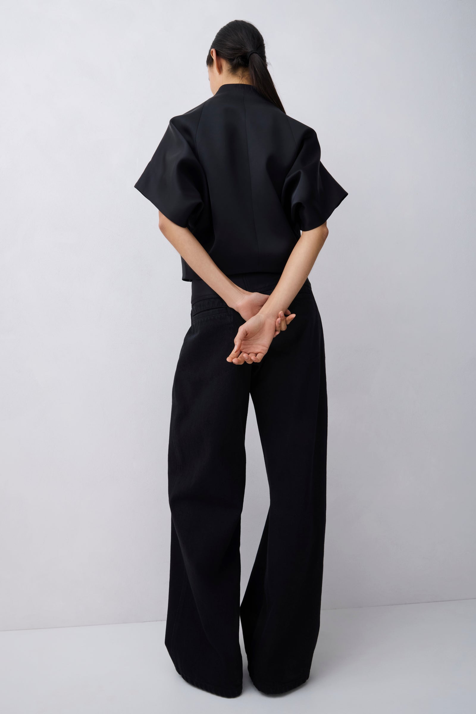 A model viewed from behind wearing a black short-sleeve top and matching wide-leg trousers against a plain light gray studio background.