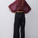 A model wearing a sheer, draped burgundy top with wide batwing sleeves and black wide-leg trousers, standing against a light gray studio background.
