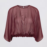 A model wearing a sheer, draped burgundy top with wide batwing sleeves and black wide-leg trousers, standing against a light gray studio background.