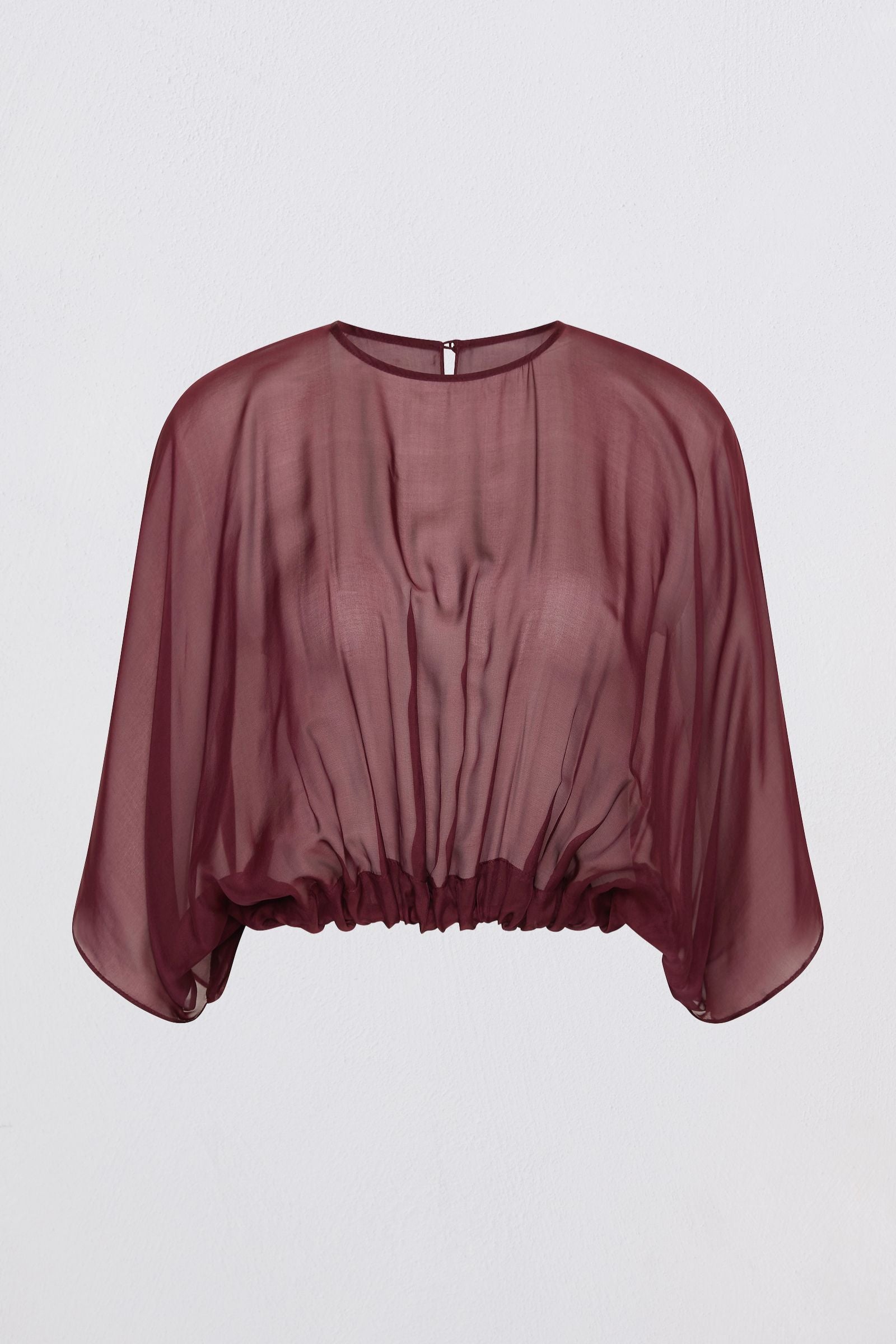 A model wearing a sheer, draped burgundy top with wide batwing sleeves and black wide-leg trousers, standing against a light gray studio background.