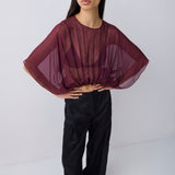 A model wearing a sheer, draped burgundy top with wide batwing sleeves and black wide-leg trousers, standing against a light gray studio background.