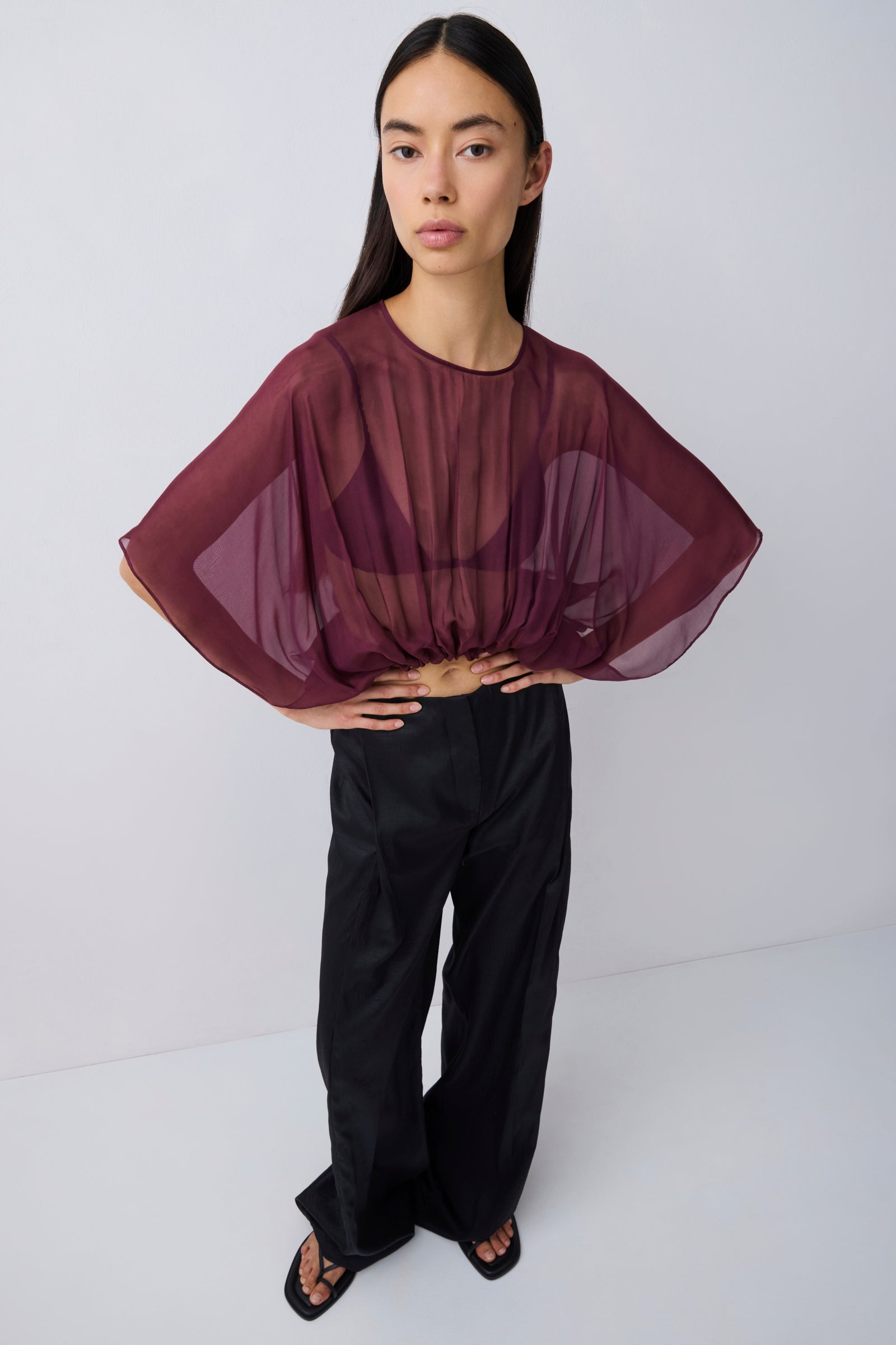 A model wearing a sheer, draped burgundy top with wide batwing sleeves and black wide-leg trousers, standing against a light gray studio background.