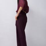 A woman modeling a dark plum purple two-piece outfit consisting of a short-sleeve draped top and a matching high-waisted maxi skirt, photographed against a plain light gray studio background.