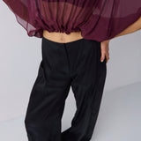 A model wearing black wide-leg trousers and black sandals, with a sheer dark purple top draped off the shoulders, shown from the waist down against a light gray studio background.