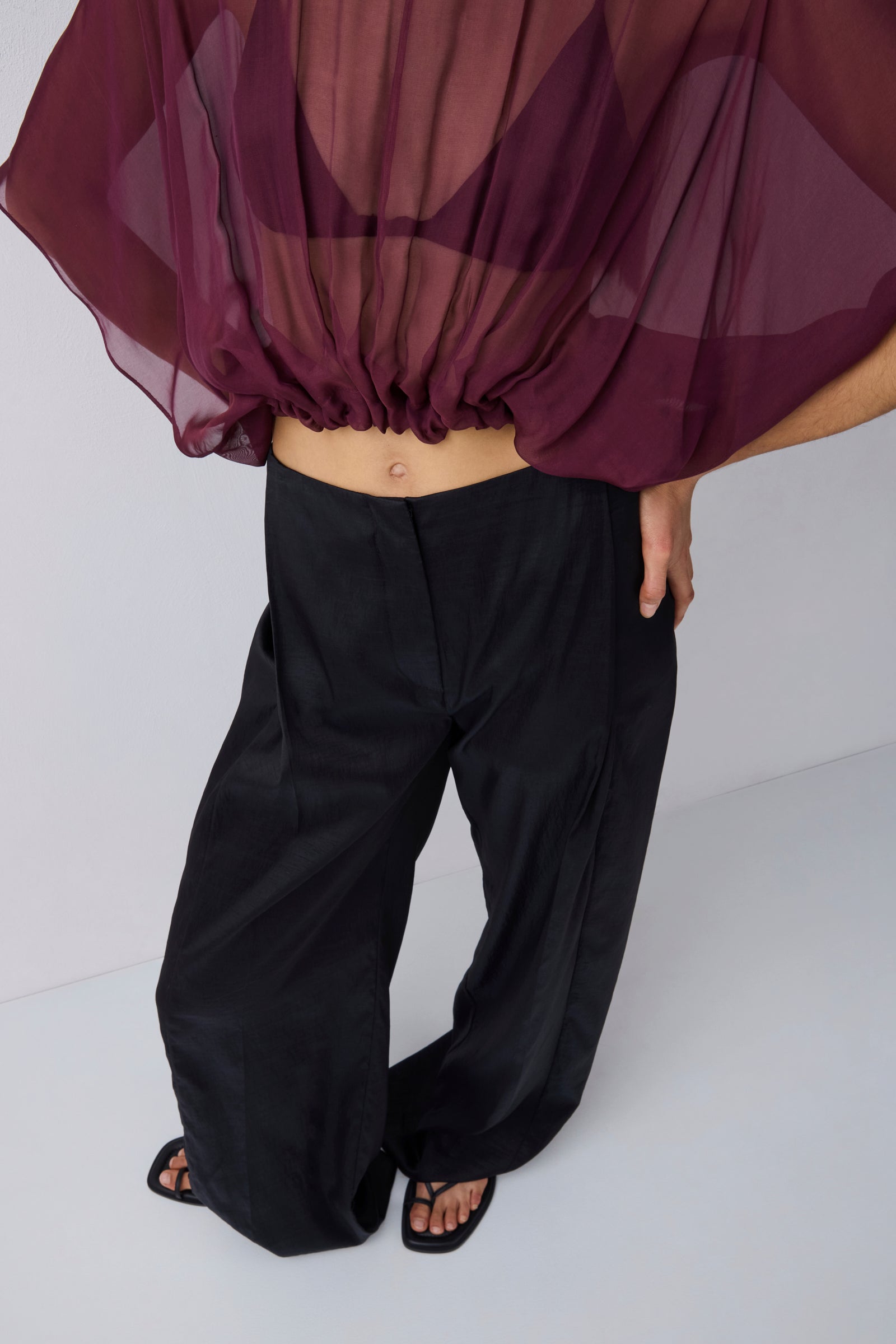 A model wearing black wide-leg trousers and black sandals, with a sheer dark purple top draped off the shoulders, shown from the waist down against a light gray studio background.