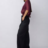 A woman modeling a sheer burgundy draped top and black wide-leg trousers against a light gray studio background.