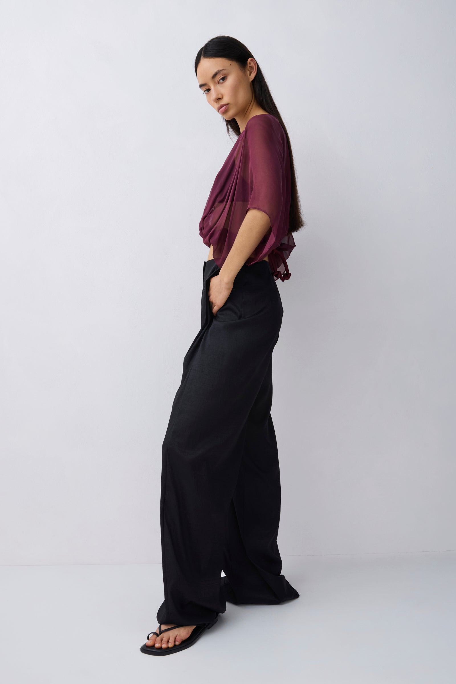 A woman modeling a sheer burgundy draped top and black wide-leg trousers against a light gray studio background.