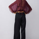 A woman modeling a sheer burgundy top with dramatic wide sleeves and a drawstring back, paired with black wide-leg pants, shown from the back against a light gray studio background.
