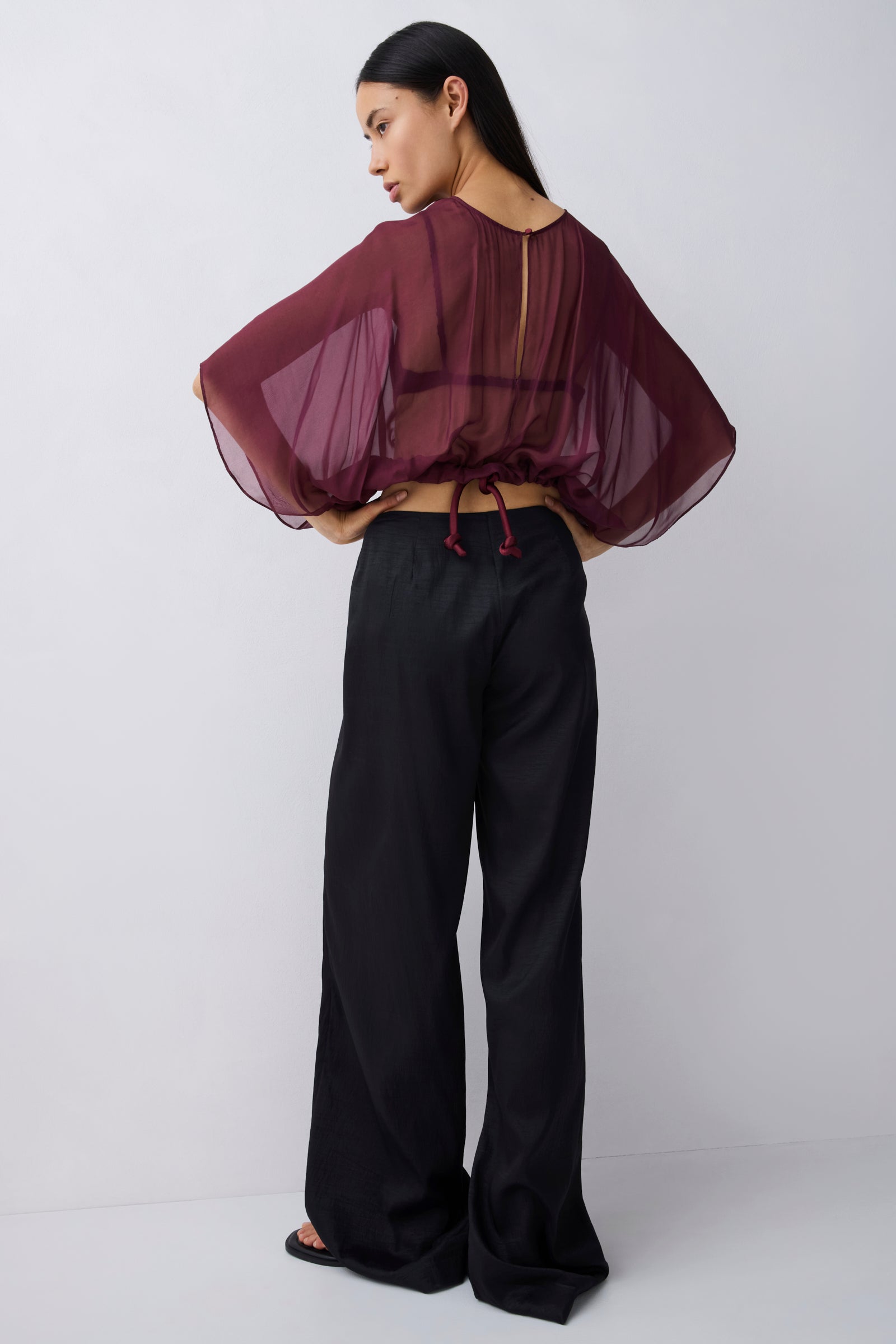 A woman modeling a sheer burgundy top with dramatic wide sleeves and a drawstring back, paired with black wide-leg pants, shown from the back against a light gray studio background.