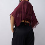 A woman wearing a sheer burgundy short-sleeve top with a tie-back detail and black wide-leg pants, photographed against a plain light gray studio background.