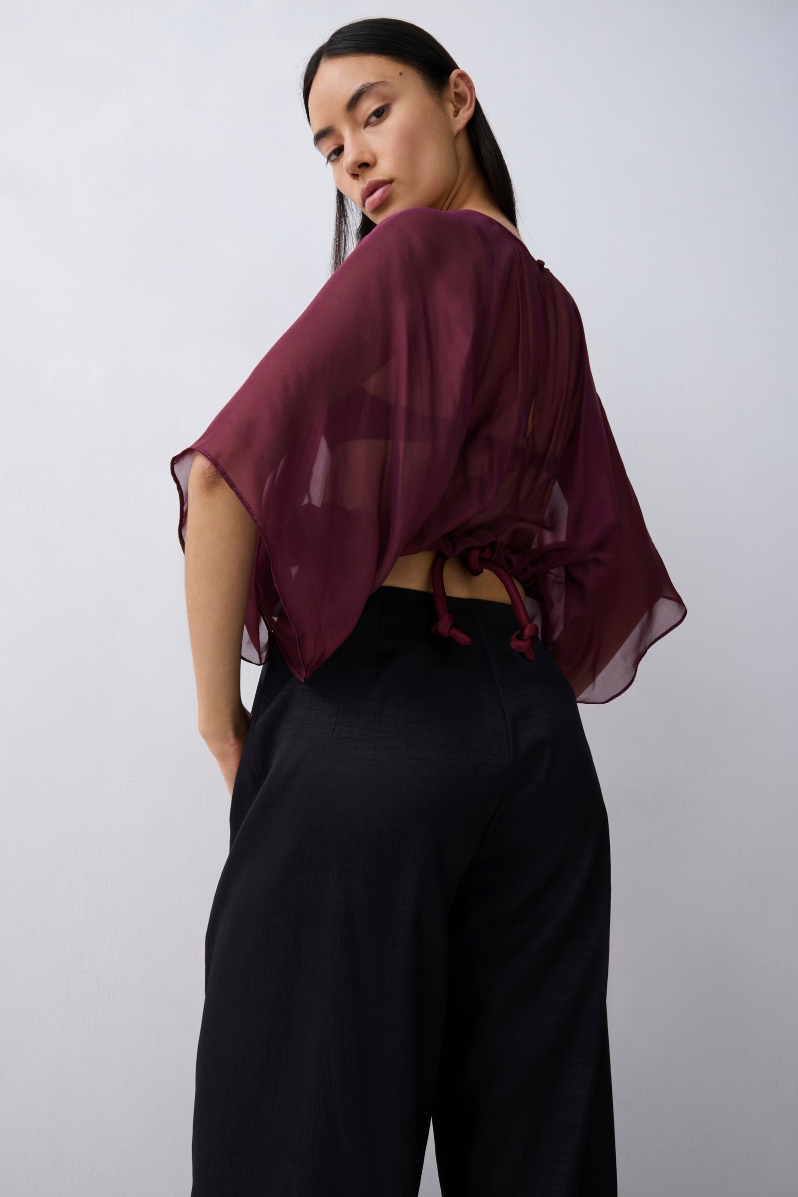 A woman wearing a sheer burgundy short-sleeve top with a tie-back detail and black wide-leg pants, photographed against a plain light gray studio background.