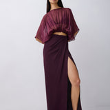 A model wearing a dark plum purple two-piece set consisting of a sheer draped short-sleeve crop top and a matching high-slit maxi skirt, paired with dark brown sandals, against a light gray studio background.