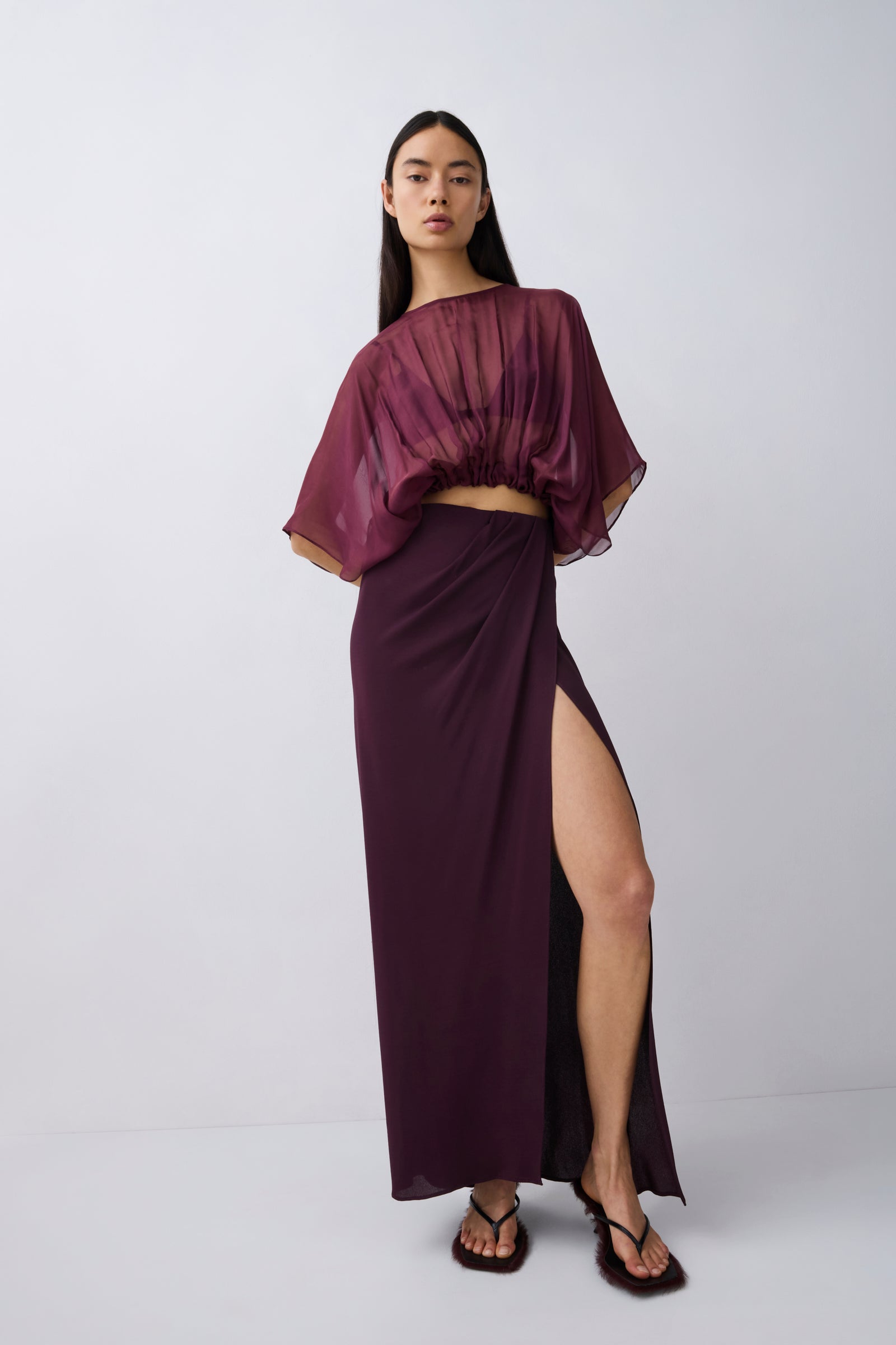 A model wearing a dark plum purple two-piece set consisting of a sheer draped short-sleeve crop top and a matching high-slit maxi skirt, paired with dark brown sandals, against a light gray studio background.