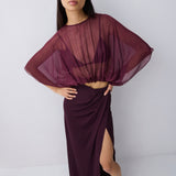 A model wearing a sheer, draped deep burgundy top with wide batwing sleeves and a matching high-slit wrap skirt, paired with black strappy heels, against a light gray studio background.