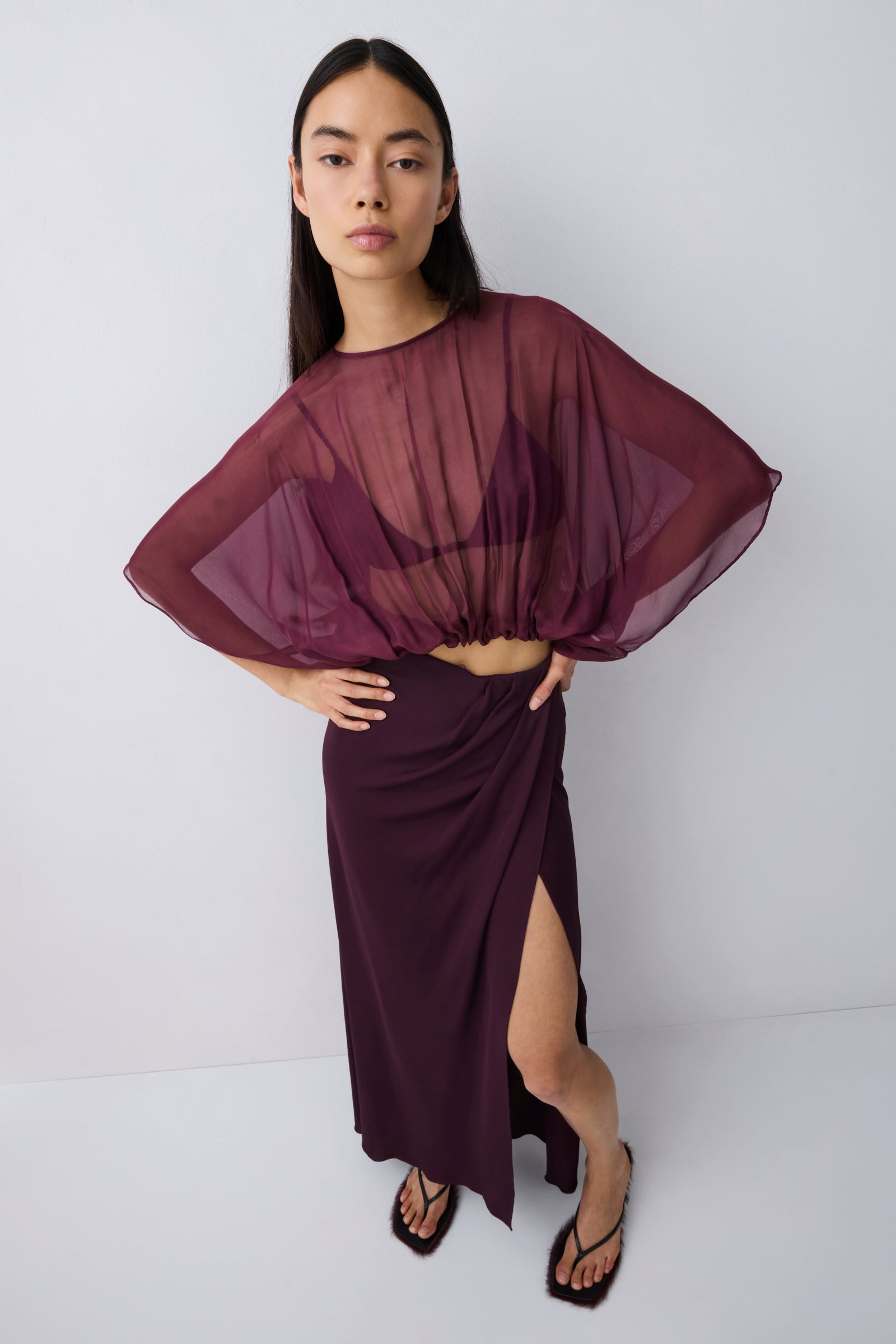 A model wearing a sheer, draped deep burgundy top with wide batwing sleeves and a matching high-slit wrap skirt, paired with black strappy heels, against a light gray studio background.