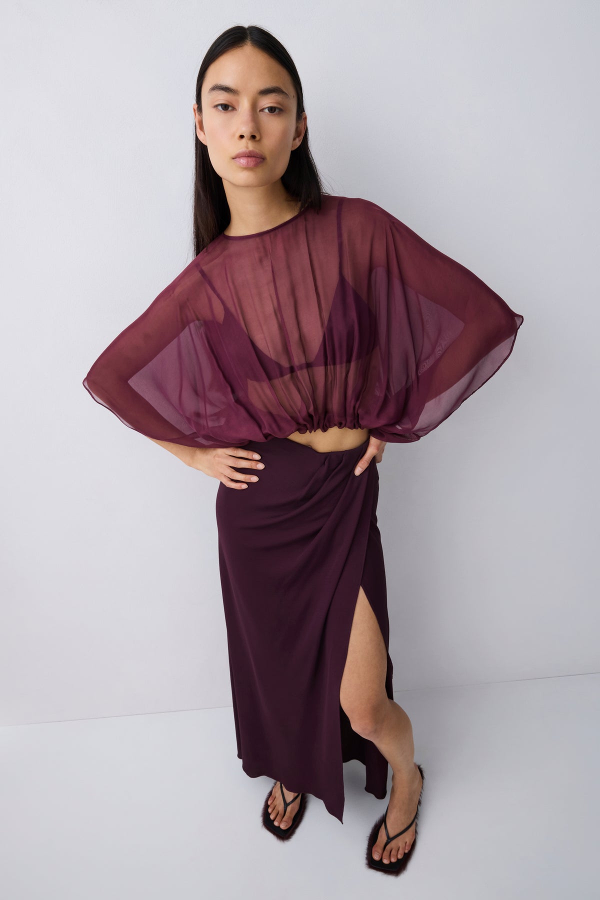 A model wearing a sheer, draped deep burgundy top with wide batwing sleeves and a matching high-slit wrap skirt, paired with black strappy heels, against a light gray studio background.