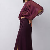 A model wearing a dark plum purple two-piece outfit consisting of a sheer draped long-sleeve crop top and a matching high-slit maxi skirt, posed against a light gray studio background.
