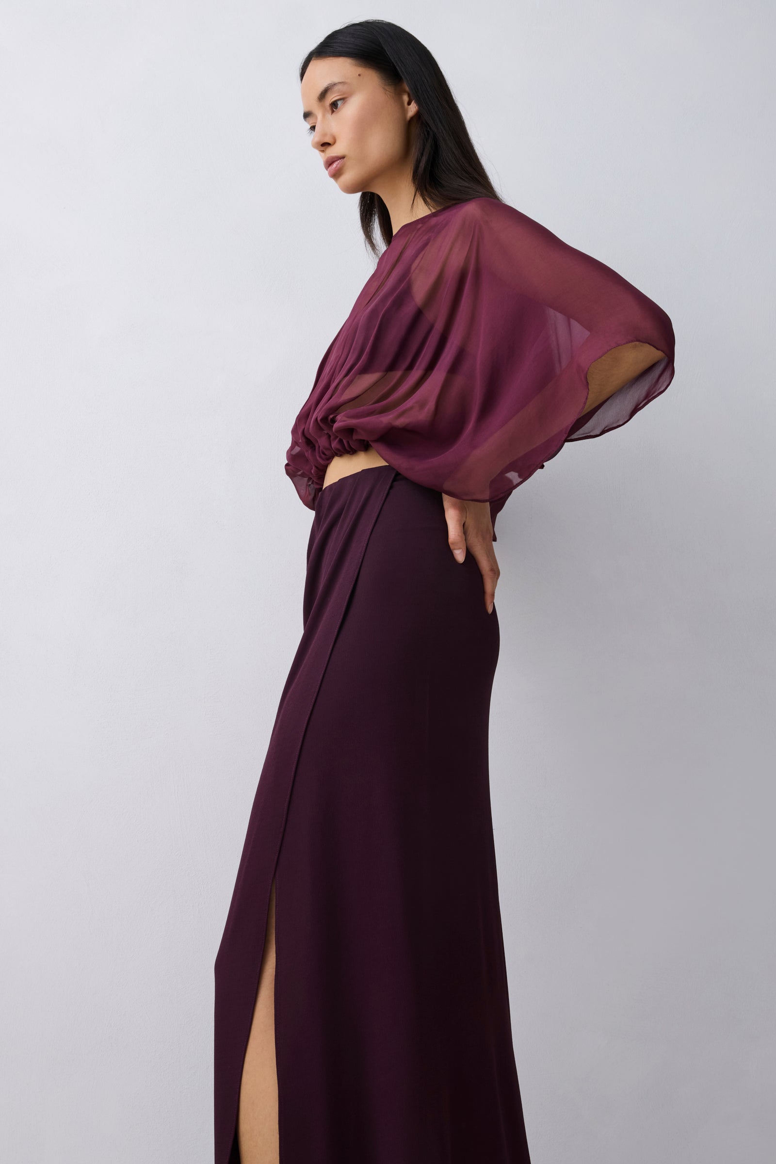 A model wearing a dark plum purple two-piece outfit consisting of a sheer draped long-sleeve crop top and a matching high-slit maxi skirt, posed against a light gray studio background.