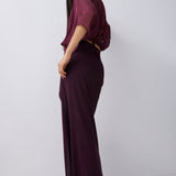 A woman modeling a dark plum purple two-piece outfit consisting of a short-sleeve draped top and a matching high-waisted maxi skirt, photographed against a plain light gray studio background.