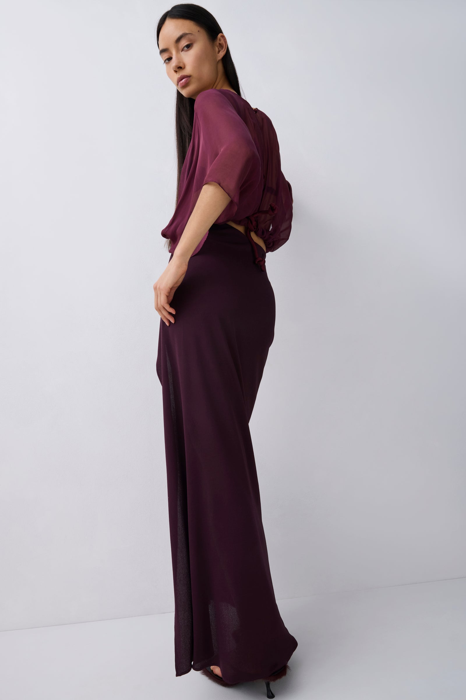 A woman modeling a dark plum purple two-piece outfit consisting of a short-sleeve draped top and a matching high-waisted maxi skirt, photographed against a plain light gray studio background.