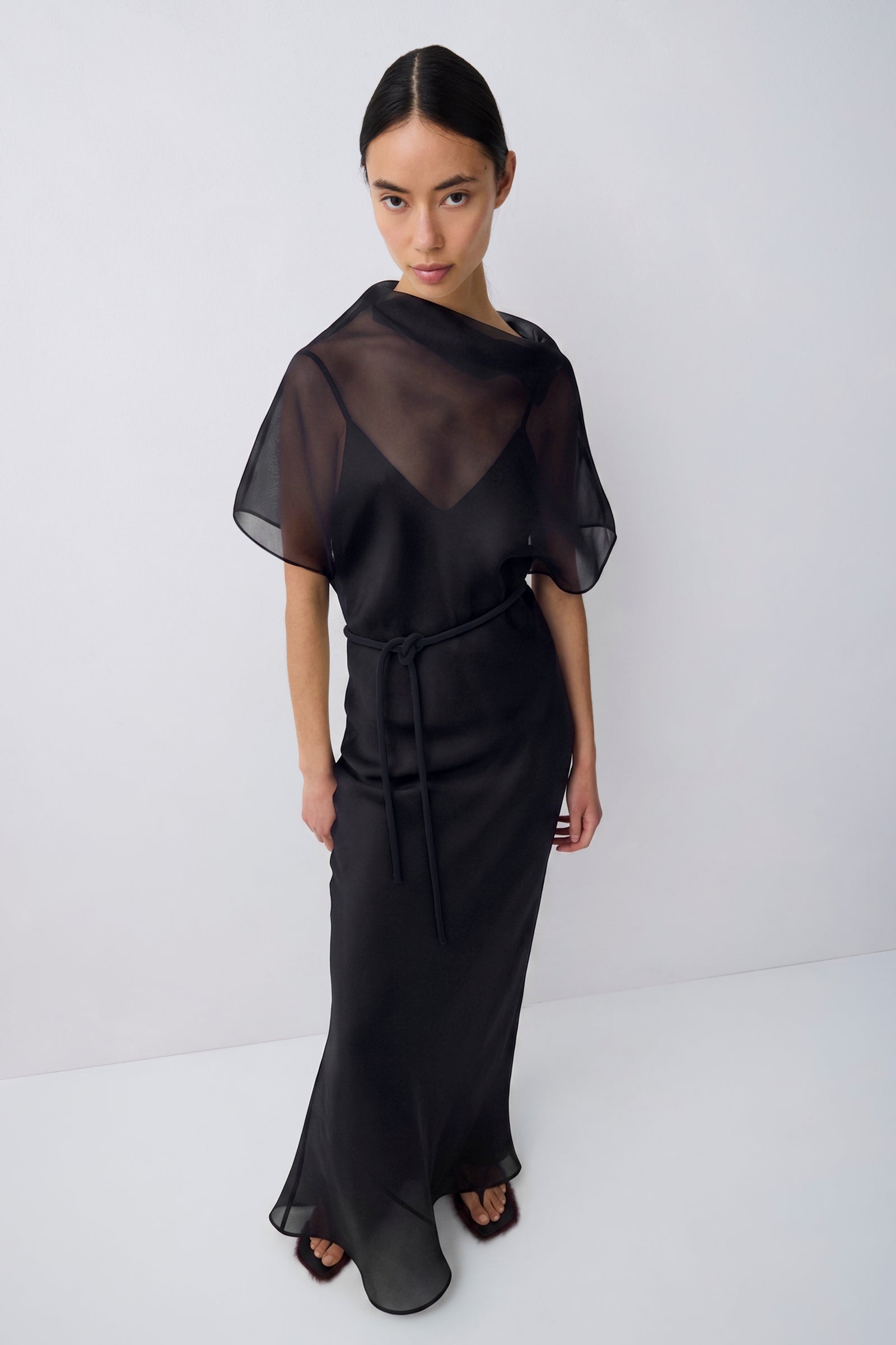 A model wearing a black sheer mesh dress with short flutter sleeves, a V-neckline, and a matching fabric tie belt at the waist, photographed against a plain light gray studio background.