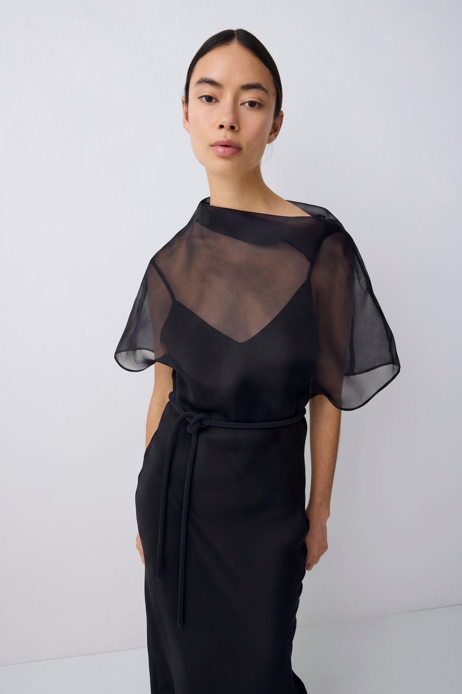 A model wearing a black midi dress with a sheer mesh yoke and flutter sleeves, standing against a light gray studio background.